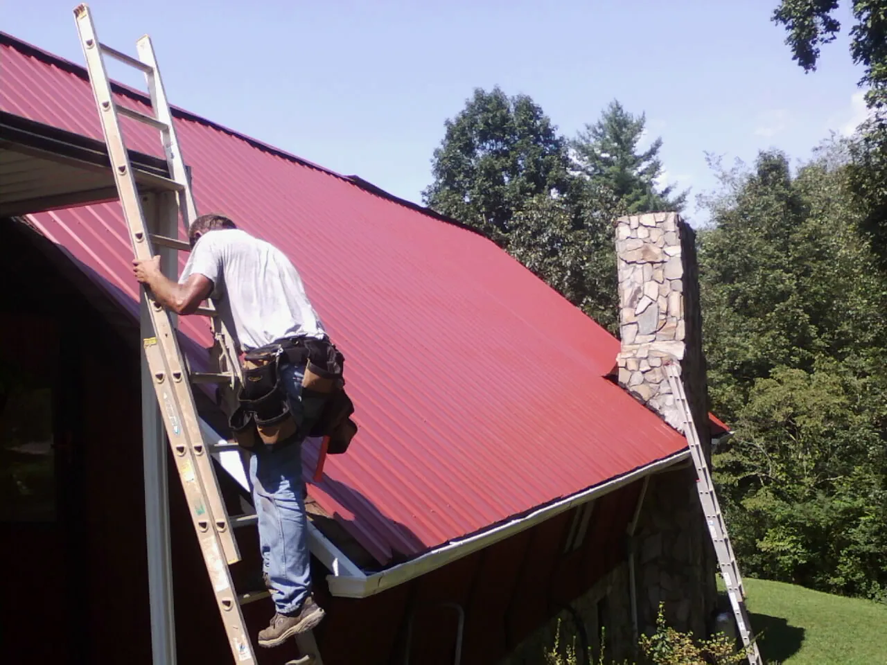 Roofer on a ladder beside a red metal roof and stone chimney — Soffit & Fascia Repair by Roofing Brothers in Aleppo